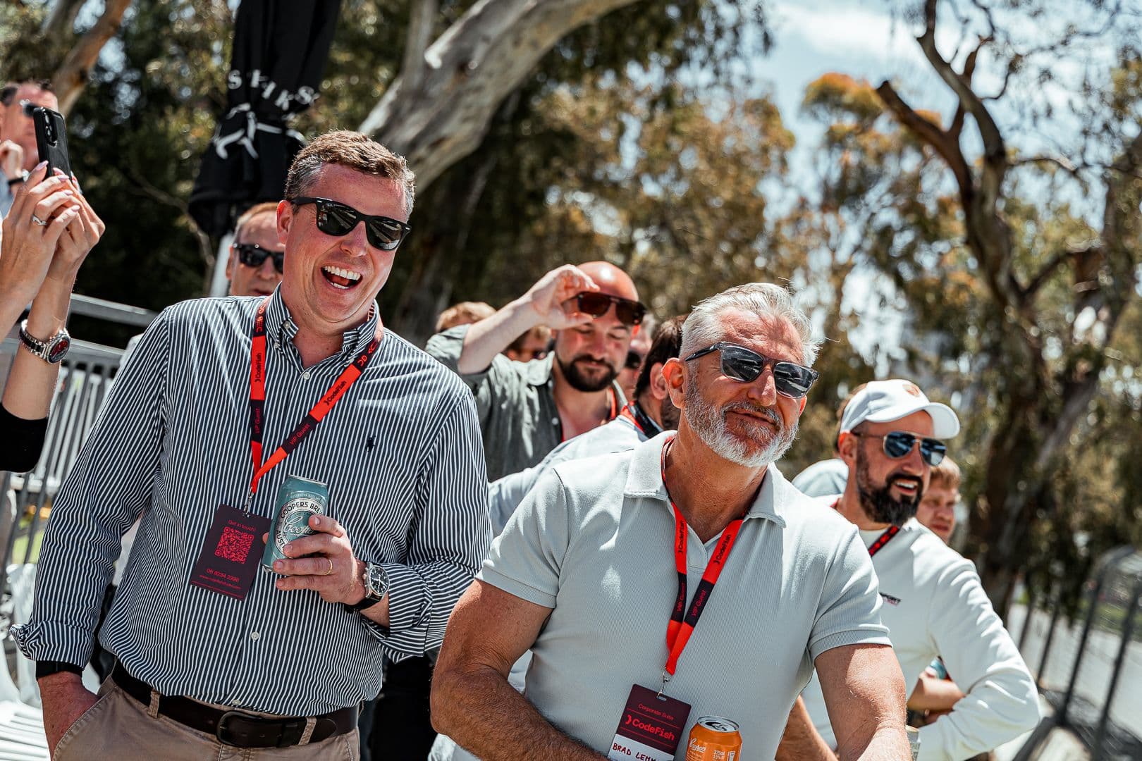 Guests in sunglasses and lanyards laughing outdoors at a trackside event