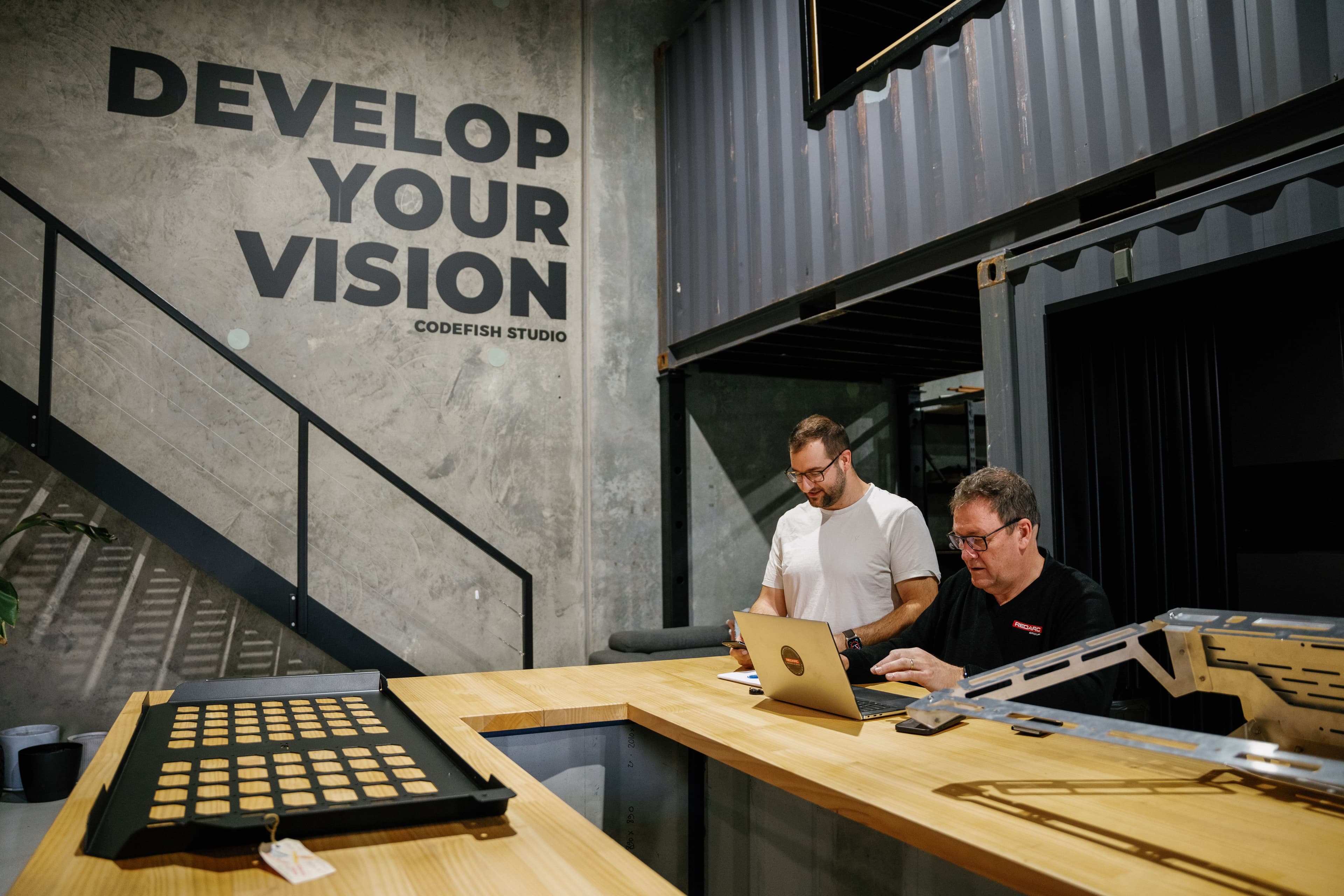 Two team members working on a laptop at a workshop bench under a Develop Your Vision wall