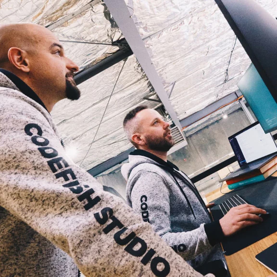 Two team members in CodeFish Studio hoodies working at a monitor in a warehouse-style office
