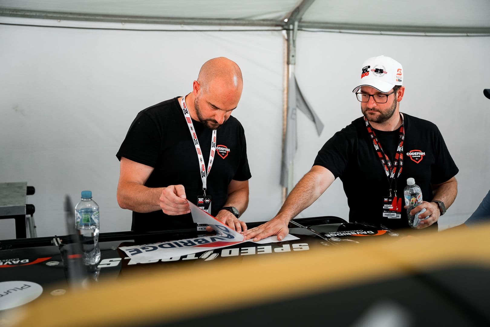 Two team members applying sponsor decals to race truck body panels in a pit marquee