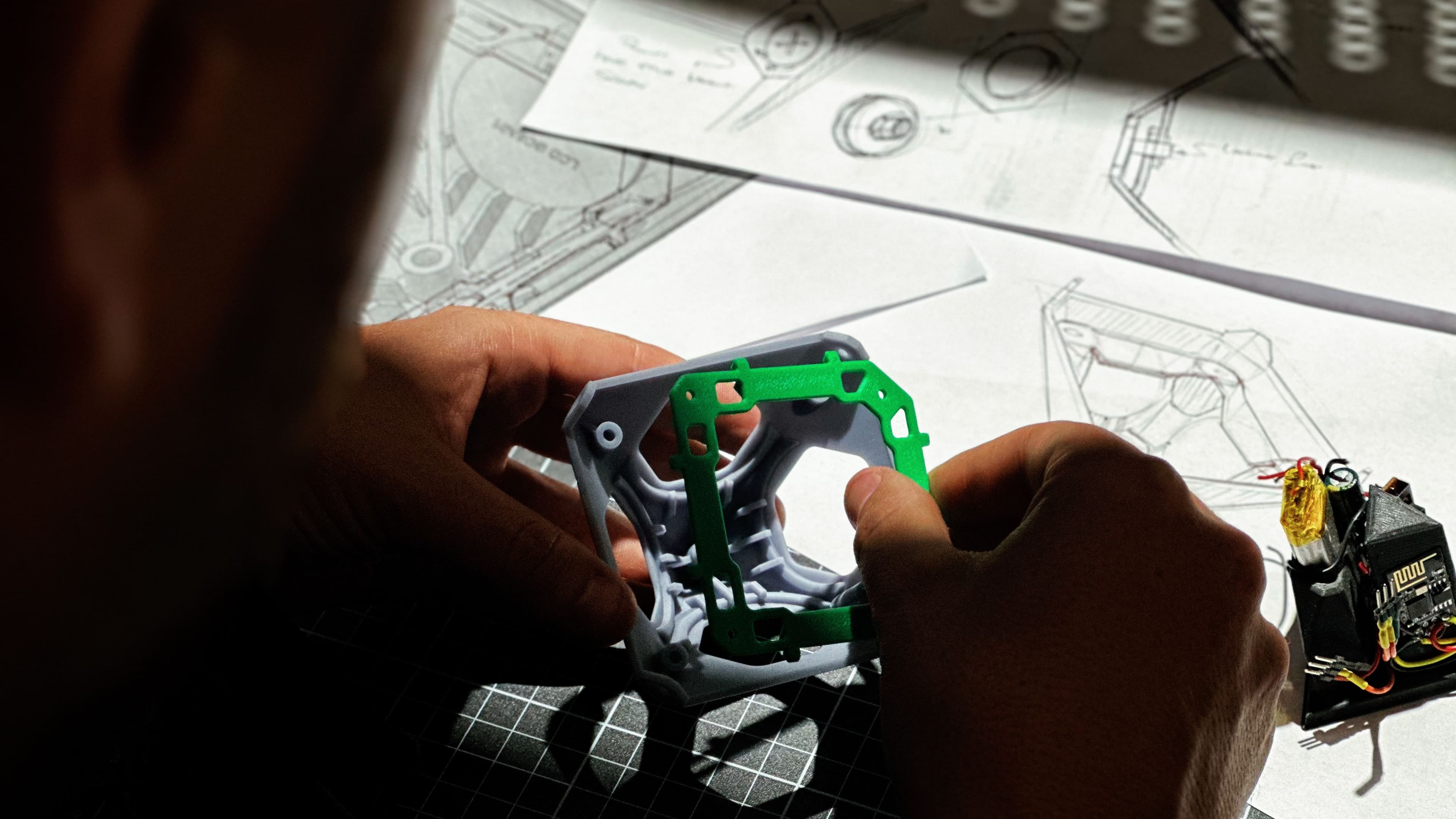 Hands holding a 3D-printed green and grey prototype part over engineering sketches